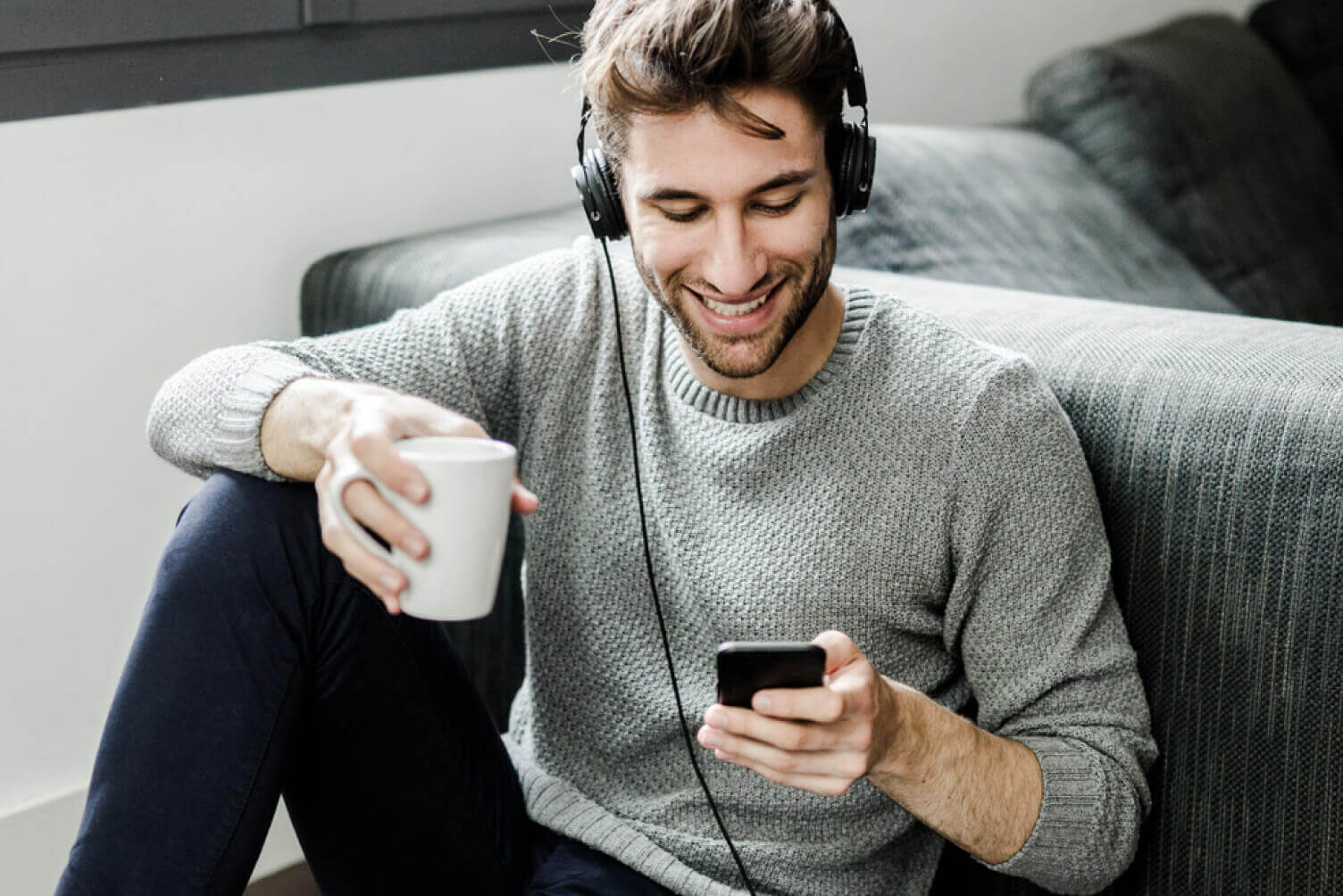 Hombre sonriendo con un café en la mano, escuchando música y viendo el celular